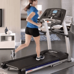 A fit woman in athletic attire running on the Life Fitness F1 Smart Treadmill in a living room setting with a television, 3 bright windows and a chair