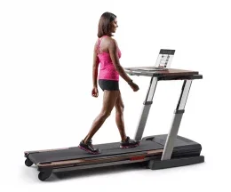 A fit woman in athletic attire walking on the NordicTrack Treadmill Desk Platinum Treadmill watching something on her laptop